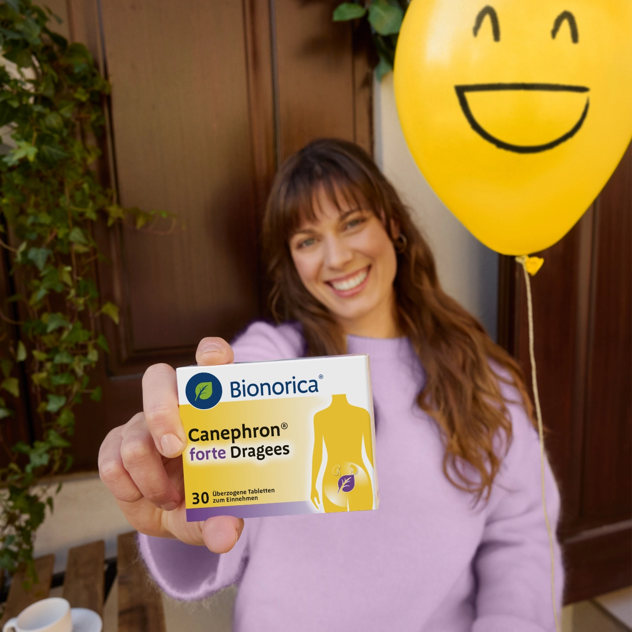 Frau hält Canephron Packshot in die Kamera, hinter ihr ein gelber lächelnder Ballon